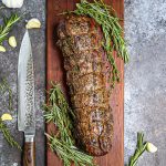 Herb Crusted Grilled Beef Tenderloin on a cutting board with herbs and garlic scattered around.