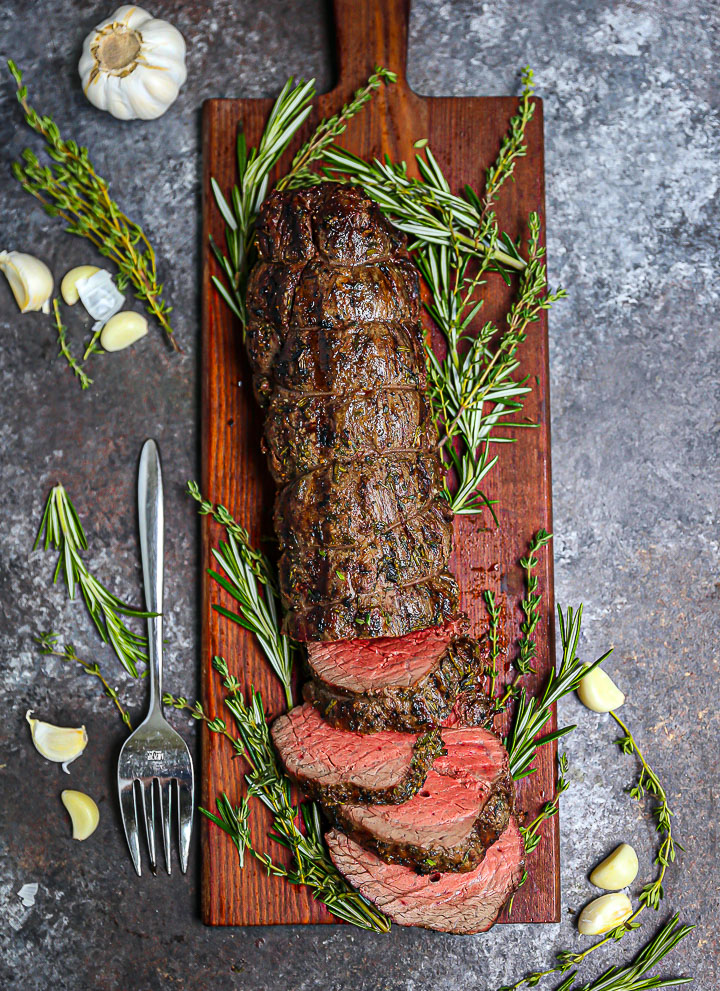 Herb Crusted Grilled Beef Tenderloin partially sliced on a cutting board with herbs and garlic scattered around.