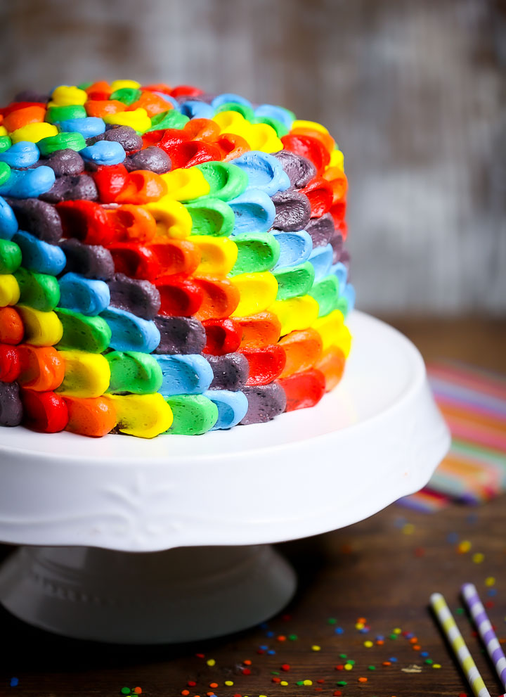 Rainbow Layer Cake on a white cake stand with rainbow napkins, sprinkles, and straws.
