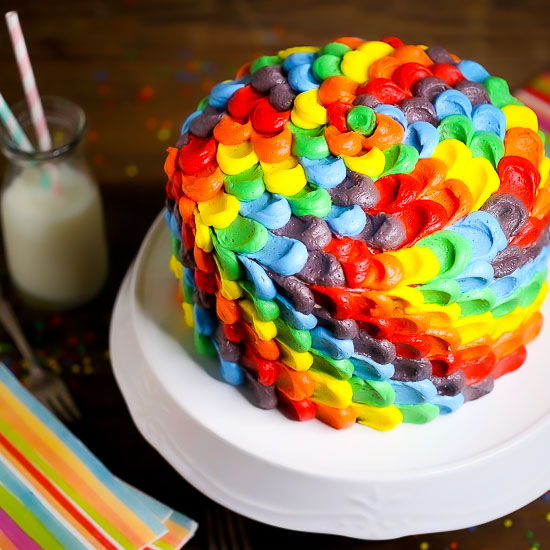 Rainbow Layer Cake on a white cake stand with rainbow napkins, sprinkles, straws and a glass of milk in the background.