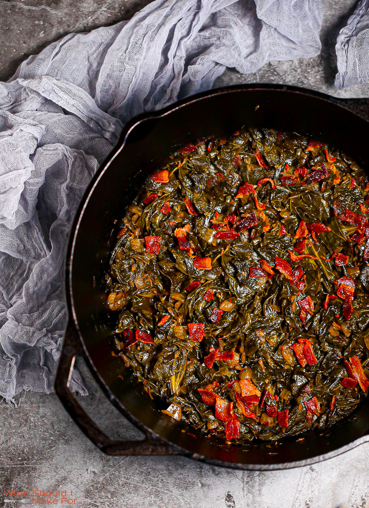 A close up shot of Braised Collard Greens with Bacon in a cast iron pot with a gray towel alongside.