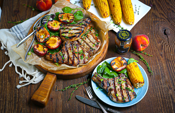 Mustard Glazed Grilled Pork Chops on a round wood board served with grilled peaches, corn, spinach, thyme and Maille mustard.