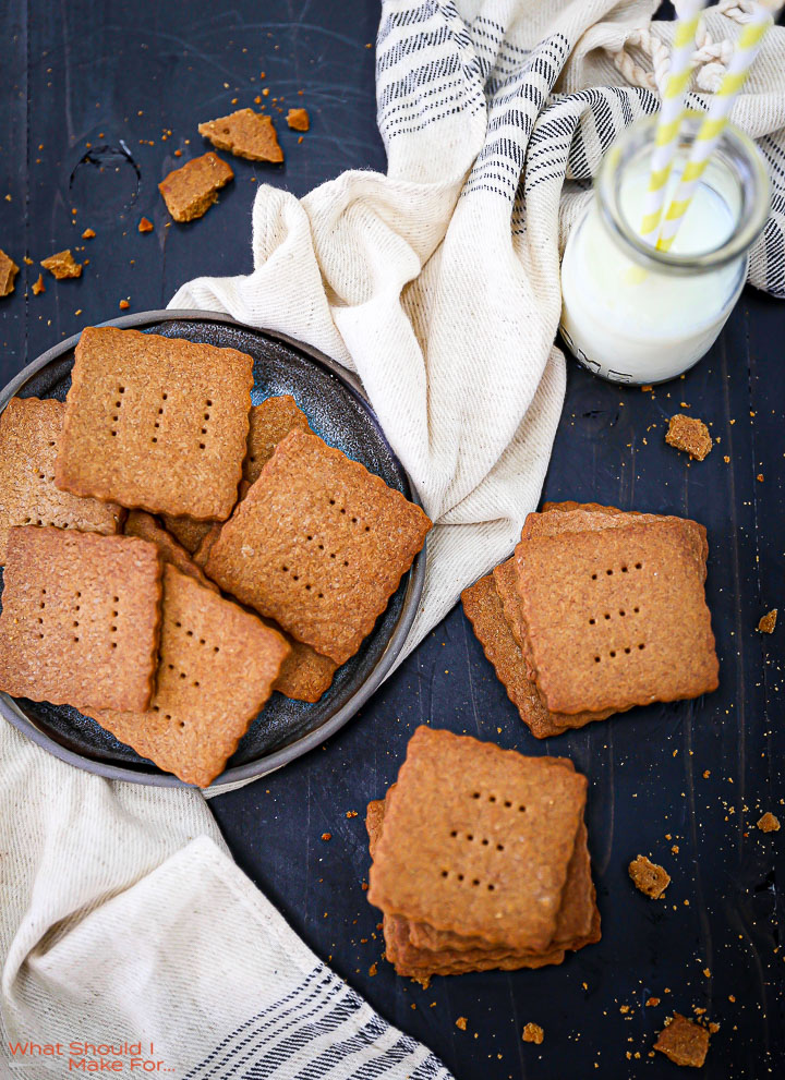 Two stacks of Homemade Graham Crackers on a black table top with crumbs scattered around and a plate of crackers and milk next to them..