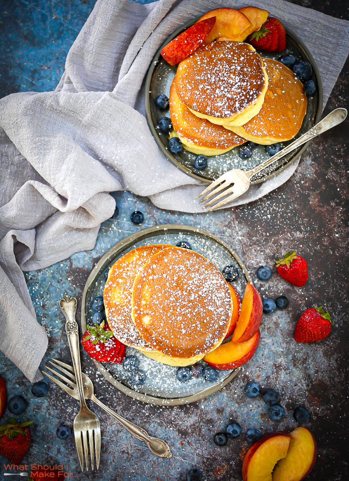 Fluffy Japanese Pancakes on two blue plates dusted with powdered sugar with peaches and berries scattered around.