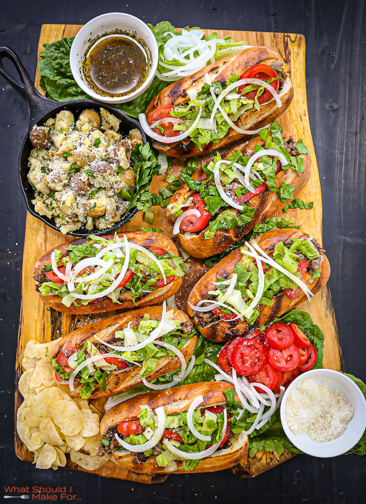 Six Cheeseburger Subs on a wooden board with a chops, sub sauce and a skillet of potatoes on it.