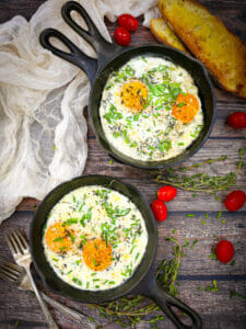 Skillet Baked Eggs sprinkled with chives and herbs, with two forks, tomatoes, toast, white table lines, and fresh thyme scattered around.
