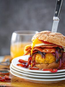 Egg Topped Bacon Cheeseburger stabbed with a knife on a stack of white plates with a glass of beer in the background.