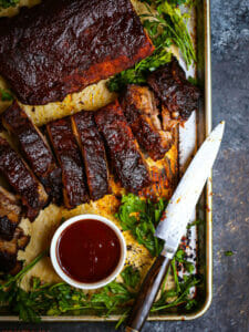 Baby Back Ribs with Bourbon BBQ Sauce on a sheet tray with herbs, sauce and cut with a knife.