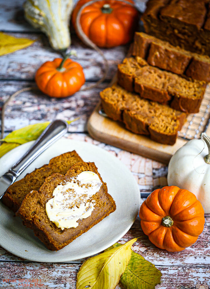 Sliced and buttered pumpkin bread on a plate with pumpkins and leaves scattered around.