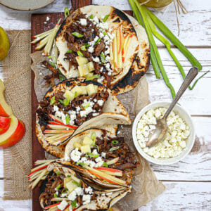 Slow Roasted Lamb Tacos on a long wooden board with apples. scallions, and feta cheese on the table.
