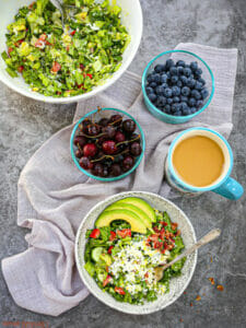 A serving of Chopped BLT Breakfast Salad on the table with coffee, cherries, blueberries and a the serving bowl of salad.