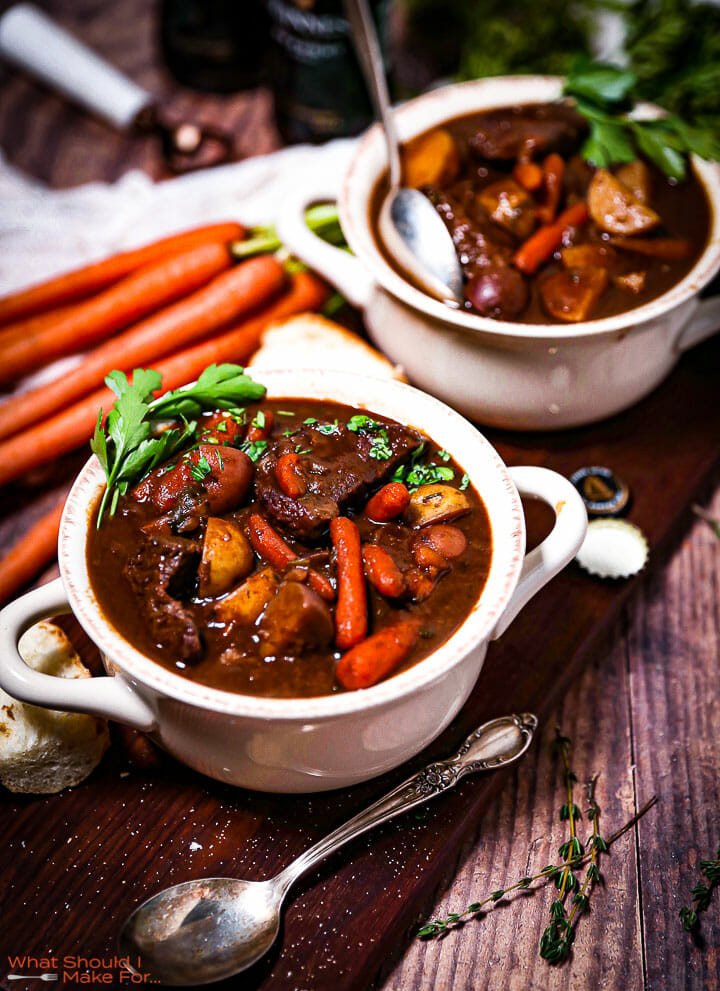 Two servings of Guinness Braised Beef Stew in crocks next to carrots and spoons.