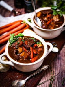 Two servings of Guinness Braised Beef Stew in crocks next to carrots and spoons.