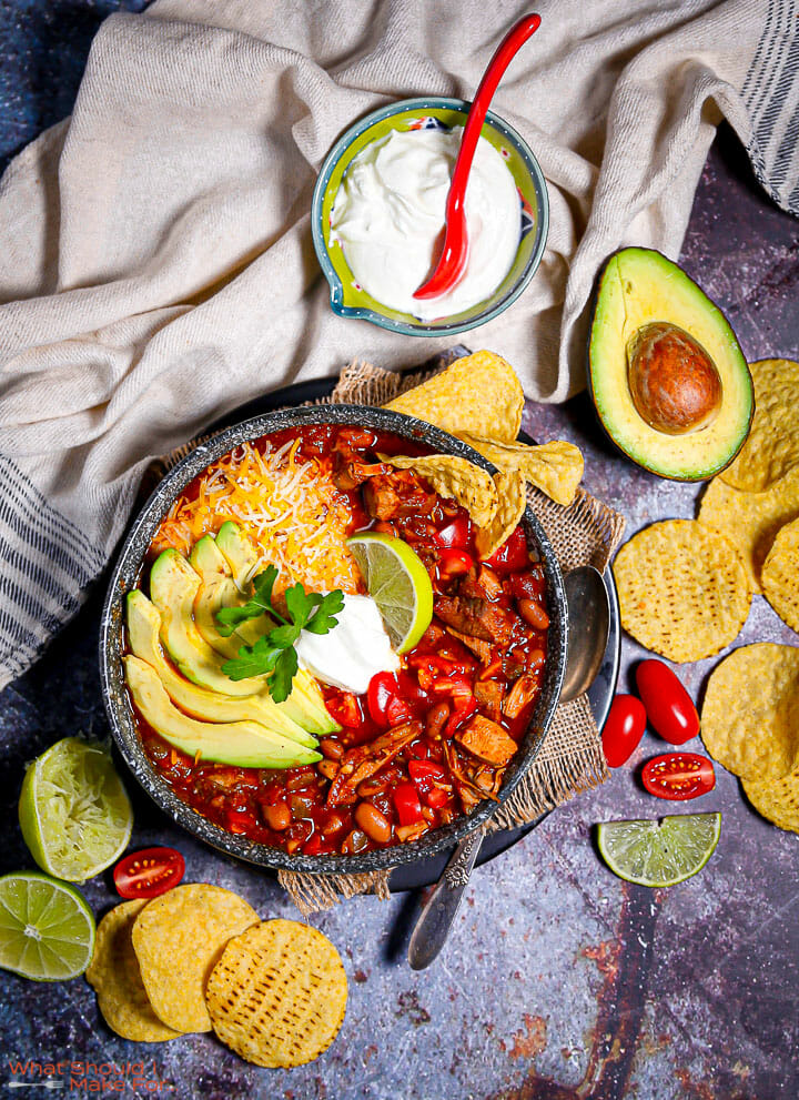 A bowl of leftover turkey chili garnished with chips, a bowl of sour cream and garnishes strewn around.