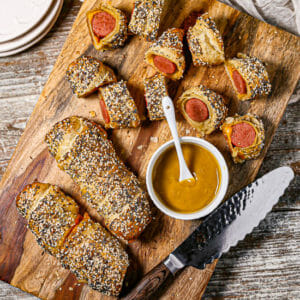 Pigs in a Blanket with a TWIST! on a wooden cutting board with mustard and a serrated knife.