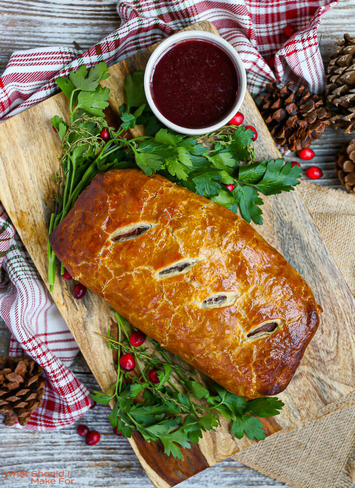 An unsliced Beef Wellington with Red Wine Sauce on a wood board with parsley and cranberries scattered around.