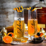 Two Bourbon Champagne Cocktails in tall flute glasses inscribed with "Celebrate" garnished with cherries and a twist of orange.