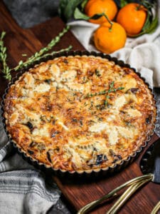 A savory mushroom tart on a wood board with clementines, fresh thyme and serving utensils next to it.