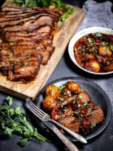 A serving of the Best Sweet and Sour Brisket on a small black plate with a knife and fork and sliced brisket in the background.