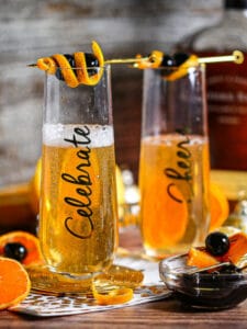 Two Bourbon Champagne Cocktails in tall flute glasses inscribed with "Celebrate" garnished with cherries and a twist of orange.