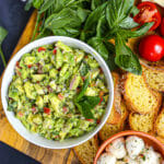 Overhead shot of Italian guacamole in a bowl with bread and basil scattered around.