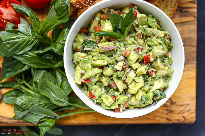 Italian guacamole in a white bowl on a wood board with basil scattered around.