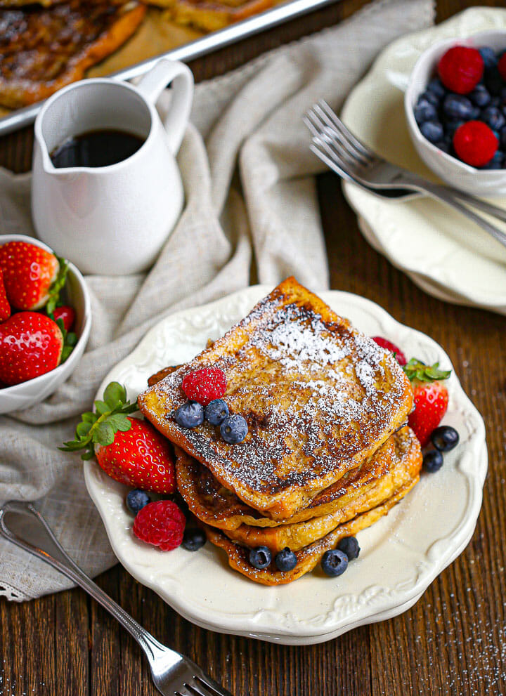 A serving of Brioche French Toast on a plate with berries and syrup in a pitcher.