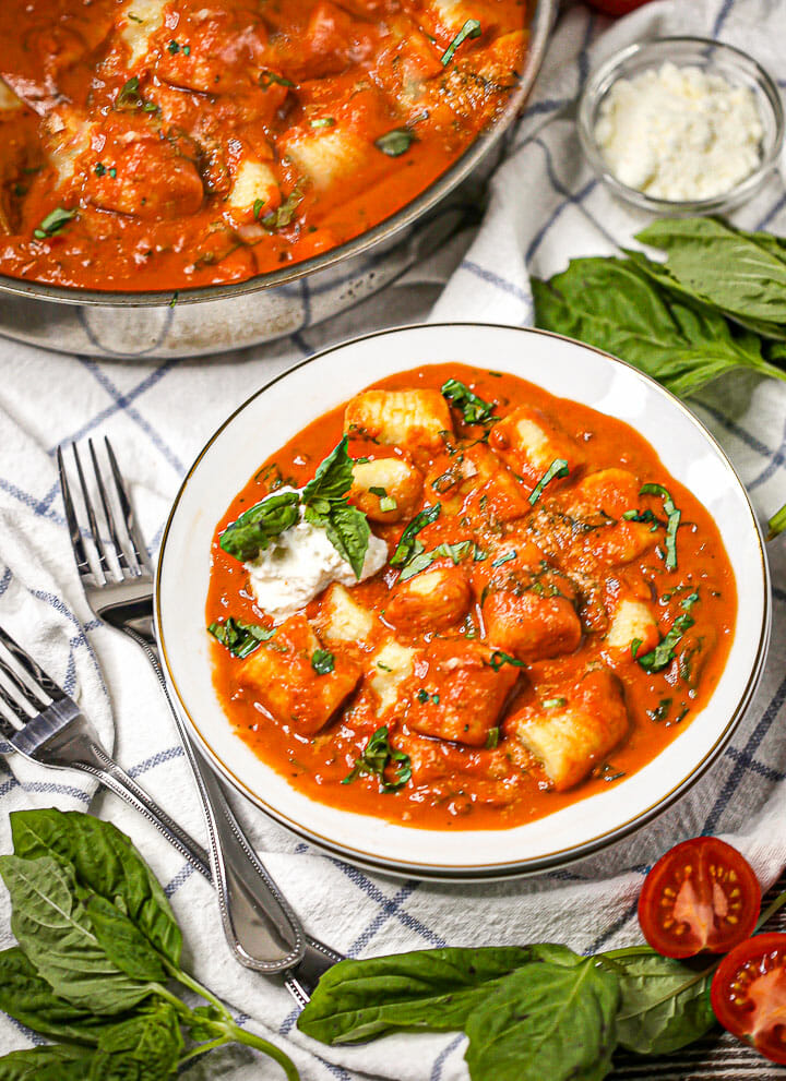 A serving of Gnocchi in Creamy Tomato Basil Sauce in a bowl with the serving dish and small dish of parm in the background.