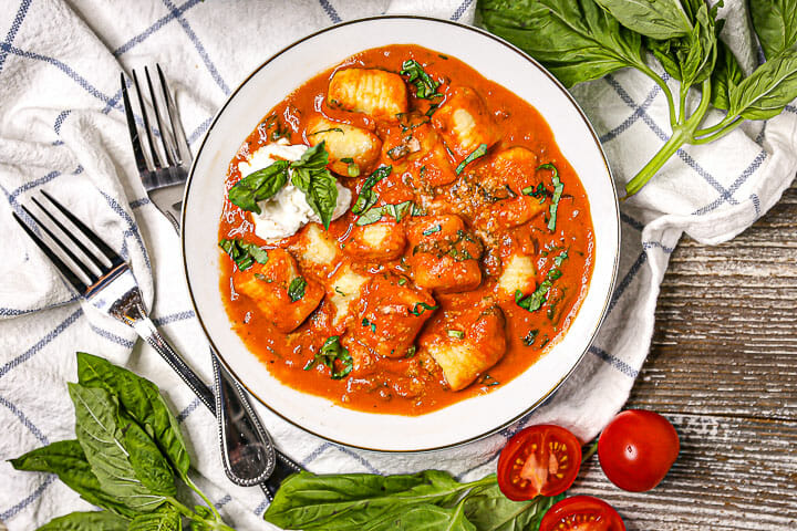 Gnocchi in Creamy Tomato Basil Sauce in a white bowl with forks, tomatoes and basil scattered around.