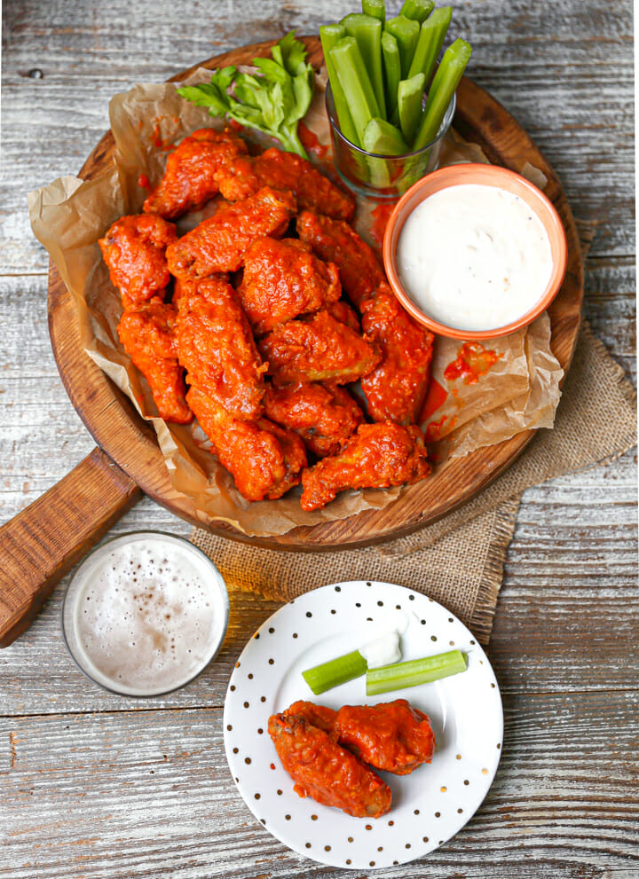 Baked Buffalo wings on a round wood serving board with celery and blue cheese and serving plate on the table too.