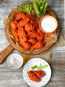 Baked Buffalo wings on a round wood serving board with celery and blue cheese and serving plate on the table too.
