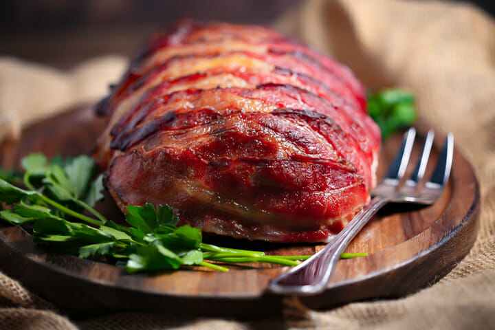 Side view of a meatloaf wrapped in bacon with a serving fork alongside.