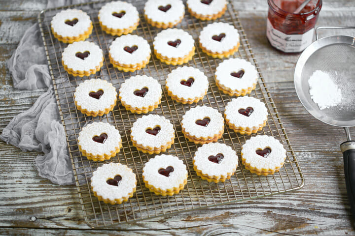 Linzer cookies on a bakers rack with a jar of jam and sifter with powdered sugar.