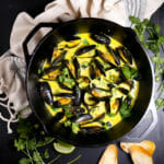 A pot of Curry Coconut Mussels with herbs and bread on scattered on the table.