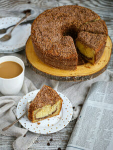A Sour Cream Coffee Cake with slices cut and one slice on a dessert plate next to a cup of coffee.