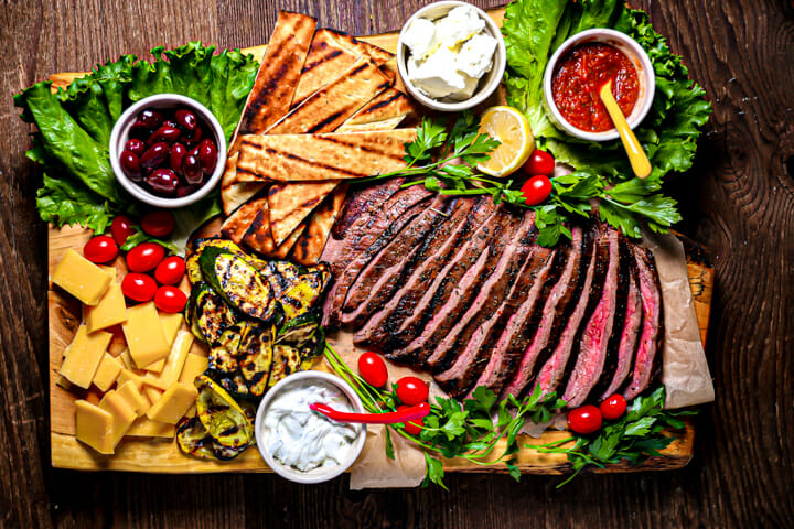 Grilled Marinated Flank Steak on a big wood board served with dips, cheese, pita and veggies.