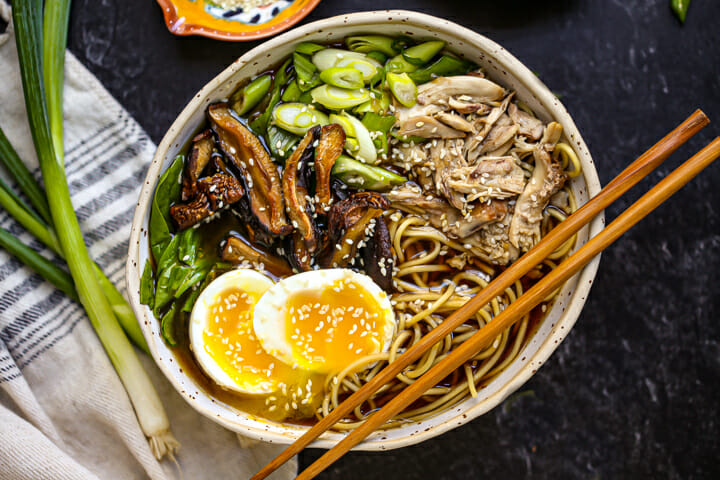 A bowl of Homemade Chicken Ramen with scallions alongside and chopsticks on the edge of the bowl.