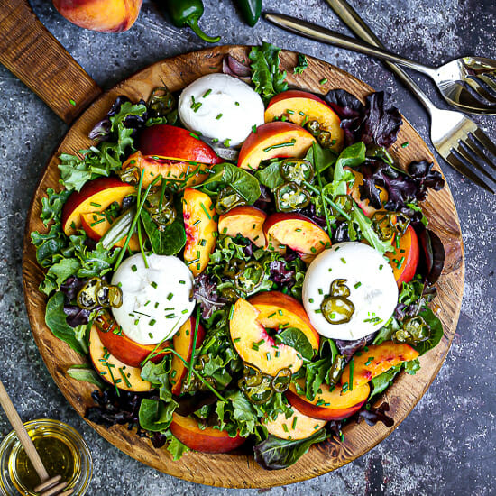 Peach and Burrata Salad on a round wood plate topped with chives and candied jalapeños.