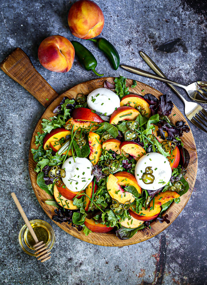 A pretty Peach and Burrata Salad with serving utensils, honey, peaches and jalapeños on the table.