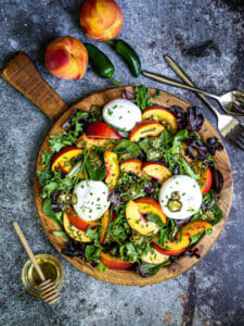 A pretty Peach and Burrata Salad with serving utensils, honey, peaches and jalapeños on the table.