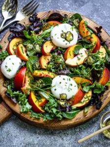 Peach and Burrata Salad served on a wood plate with honey to drizzle and peaches and jalapeños on the table.