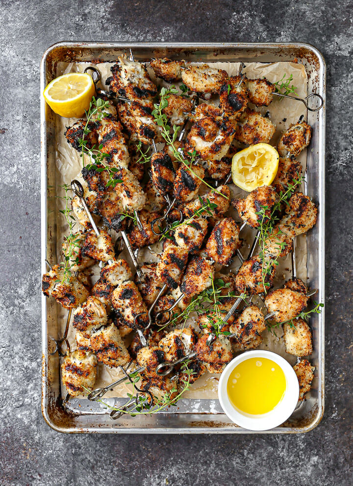 Breaded chicken skewers fresh off the grill on a sheet pan scattered with lemon and herbs.