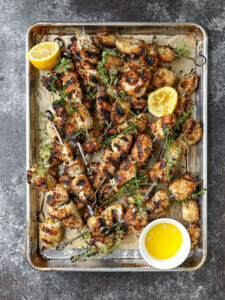 Breaded chicken skewers fresh off the grill on a sheet pan scattered with lemon and herbs.