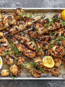 Breaded chicken skewers fresh off the grill on a sheet pan scattered with lemon and herbs.