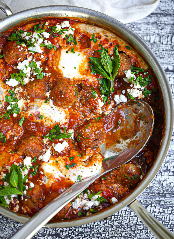 A serving spoon laying in a pan of Shakshuka with Lamb Meatballs.