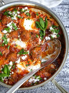 A serving spoon laying in a pan of Shakshuka with Lamb Meatballs.