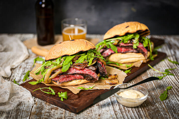 Two steak sandwiched on a wood board with a side of aioli on the table.