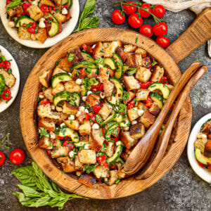 Garden Vegetable and Pancetta Panzanella on a round wood serving tray with tomatoes and basil scattered around.