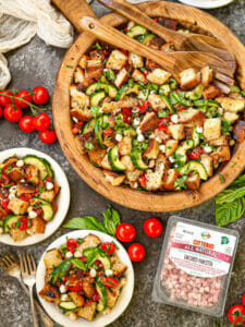 Overhead shot of panzanella salad on a wood tray with two plated servings and a package of cubetti on the table.