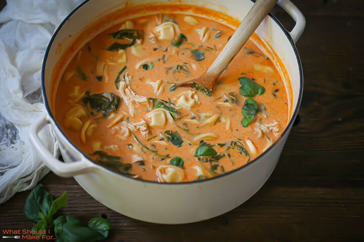 A pot of simmering Creamy Chicken Tortellini Soup with fresh basil stirred in with a wooden spoon.